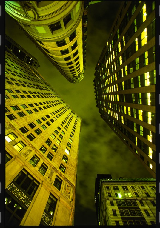 A 35mm film scan looking up between city towers at night in the NYC Financial District, sodium-lit yellow-green against a dark sky, printed with the sprocket holes left in down both edges.